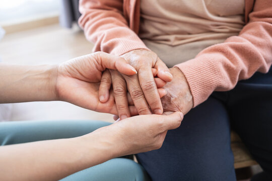 Daughter holding senior mother hand for comfort and support at home. Close up elderly care and medical empathy concept