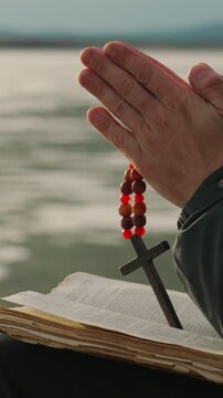 The hands of a man with a Christian cross reading the Bible on the shore of an icy lake. A man alone studies the word of God in search of truth and spirituality. Vertical video.