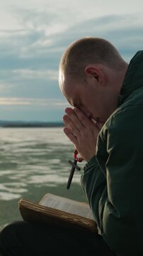 A lonely young man reads the Bible and prays to the Lord God on the shore of a lake. He holds a Christian cross in his hands. A religious concept. Vertical video.
