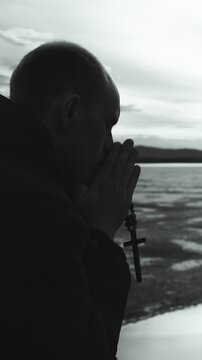 A religious young man reads the Bible and prays to the Lord God on the shore of a lake. He holds a Christian cross in his hands. Black and white. Vertical video.