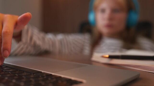 Close-up of a female student's hand using a laptop trackpad for online learning and research, with her blurred face and headphones visible in the background for a modern education concept