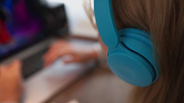 Back view of young girl with headphones studying online, doing homework on laptop. Focused student using computer for elearning, distance education, or attending virtual classroom at home
