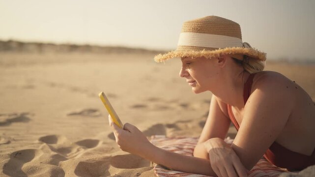 Female tourist relaxing on sunny beach in swimsuit and straw hat, checking smartphone while enjoying summer vacation, seaside leisure and carefree travel lifestyle.