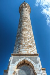 Naklejka premium Majestic soaring stone minaret with ornate carved arch, geometric tile panel and bright blue sky conveying awe