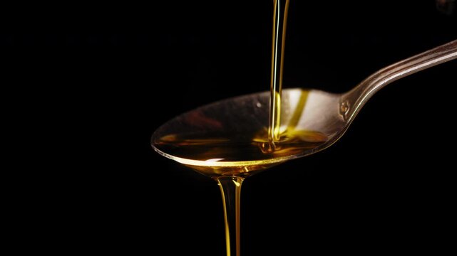 Macro shot of golden liquid olive oil pouring onto a spoon over black. Perfect cooking oil pouring into a spoon in slow motion.
