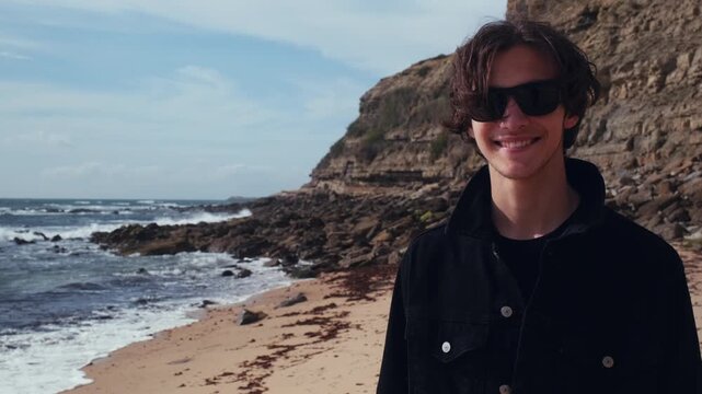 Happy, gorgeous man walks down the beach and glances at the camera. Smiling young brown-haired man with rocks and ocean in the background.  An attractive guy in sunglasses walks through nature.