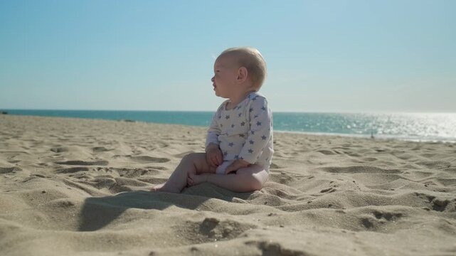 Active, cheerful toddler plays along a sandy sea beach on sunny summer day. Slow motion: Baby is playing on the sea beach on sunny day. Little boy exploring the world on sand beach against  a sea