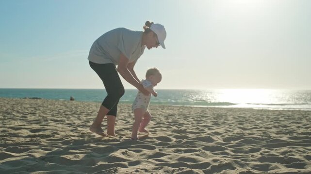 Mom teaches her baby to walk on a sandy sea beach. Toddler takes the first steps with caring mother. Kid takes first steps with the support of his mother on nature. Baby is learning to walk, outdoors.