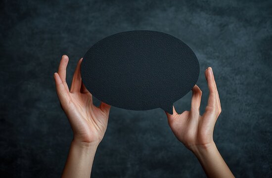 two hands holding a blank black speech bubble cutout against a dark textured background suggesting silence and invitation to speak