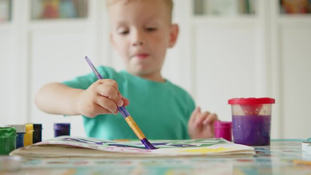 Happy child learning to draw with a brush and paint. Cute blonde boy sitting at a desk and painting with a paintbrush and gouache colors on paper at home. 