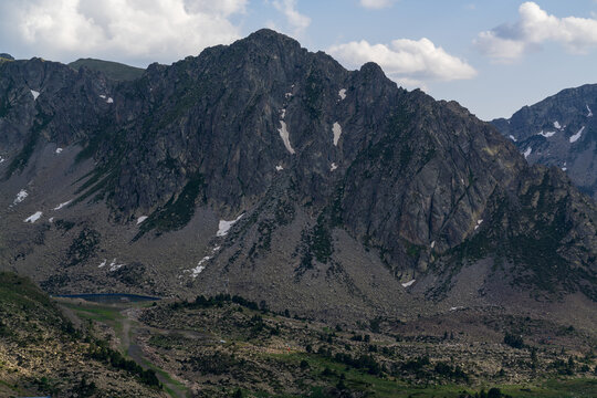 A rugged granite peak rises sharply above vast scree slopes and lingering patches of snow in the Andorran Pyrenees. Below, a small alpine lake reflects the summer sky near a winding mountain trail
