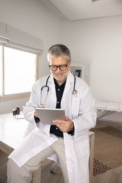 Senior male doctor in white lab coat working online on digital tablet in clinic office. Telemedicine application use, digital health records management, and professional medical consultation concept