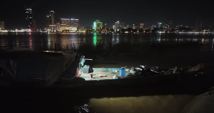 Night view of a traditional fishing boat moored on the Mekong River, lit by an interior light, with the Phnom Penh skyline in the background.