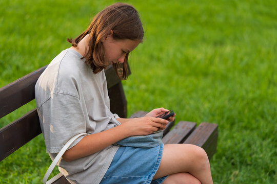 Teenage girl focuses on her phone while sitting on a bench. Dressed in casual summer attire, she is engrossed in her device. The background features lush green grass.