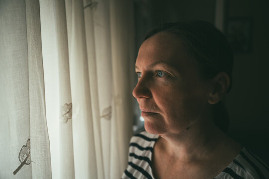 Pensive adult woman standing indoors by a window in soft daylight. Concept of solitude, mental health, contemplation, and peaceful moments at home.