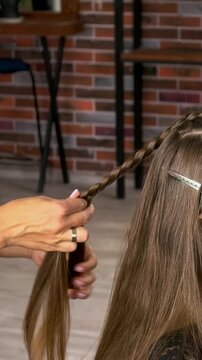 Hair stylist braids a teenage girl's long brown hair in a beauty salon, professional hairstyling, youthful fashion and creative hair care.