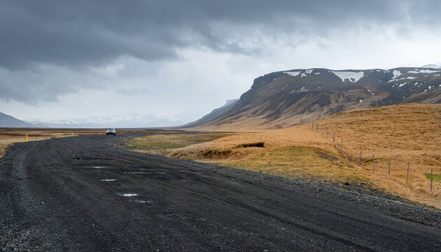 Vehicle driving along an open F249 road through vast golden-yellow fields next to snow-dusted mountains under dramatic clouds in Iceland.. Icelandic landscape