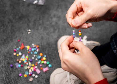 Close-up shot of a person's hands stringing colorful small beads onto a thread, with a scattered pile of beads nearby on a dark surface.