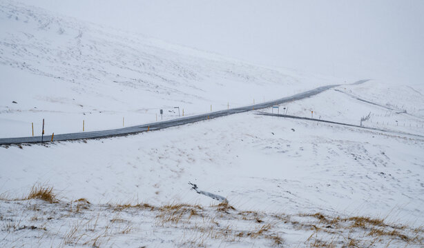 A high-altitude road cutting through a completely snow-covered mountain landscape under a thick white sky in Iceland. Snow storm Iceland