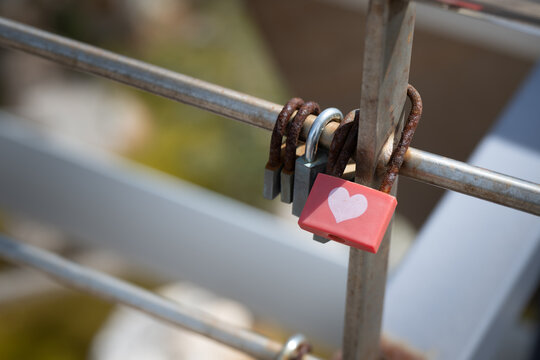 Close-up shot of several love padlocks, including a bright red lock with a white heart symbol, secured to the metal bars of a bridge railing. Romance lovers, couple, love for ever