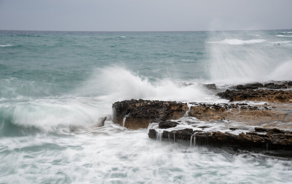 A long-exposure coastal landscape shot of powerful ocean waves crashing and splashing violently against dark, rugged rocks along the shoreline. Cape Greko peninsula Cyprus