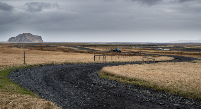 A curved black F249 gravel road leading through flat grasslands toward a distant solitary mountain peak under a moody, overcast sky in Iceland.