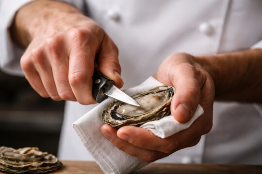 Chef's hands skillfully shucking an oyster with a knife, holding it on a cloth, wooden table and additional oysters visible in the background.