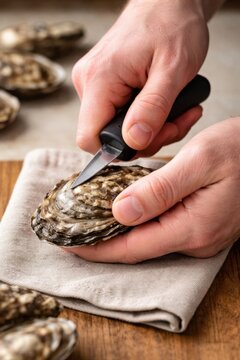 Chef's hands skillfully shucking an oyster with a knife, holding it on a cloth, wooden table and additional oysters visible in the background.