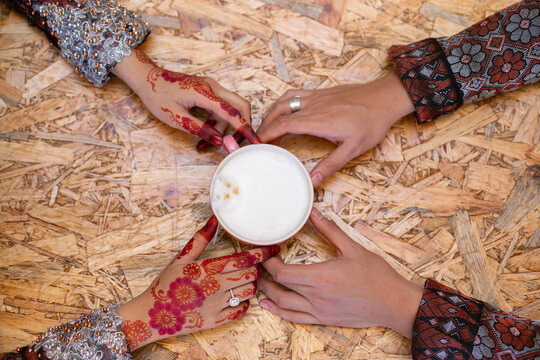Overhead flat lay of a Malay bride with intricate henna and her groom holding a coffee cup together over a textured wooden table.