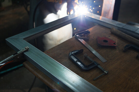 Metal welding spark beside aluminum frame and woodworking bench with clamp, ruler, and hand tool, creating focused workshop scene with dramatic light and craftsmanship