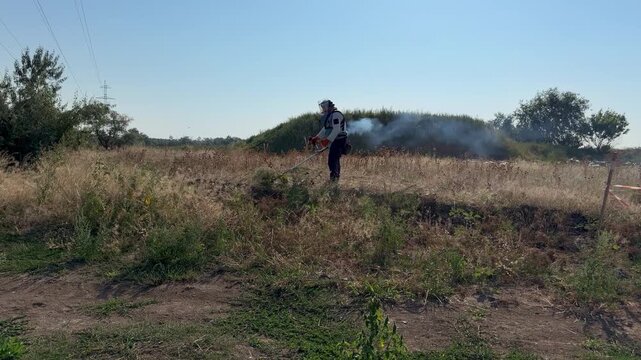 Deminer in protective visor and body armor cuts dry overgrown grass with a brush cutter on a safe strip during humanitarian mine clearance, warning tape marking the perimeter