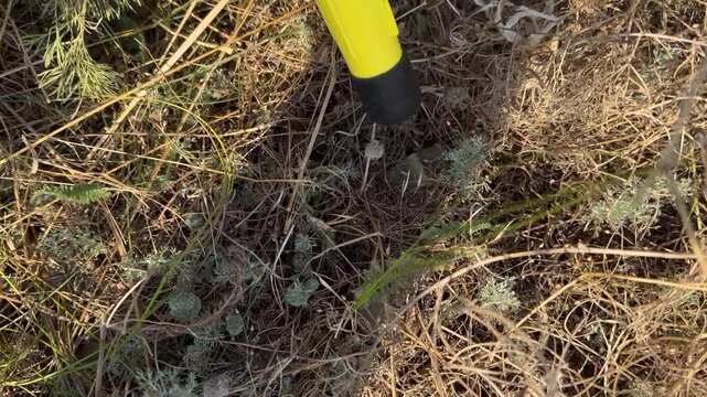 Hazardous find during humanitarian demining: parachute ribbon and metal glint of a KOBE 3B30 dual-purpose cluster submunition concealed in dry grass, marked by a yellow pointer