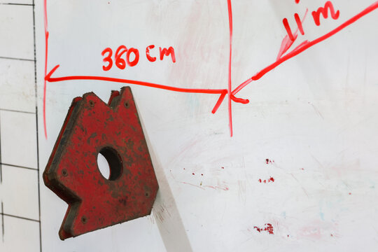Red metal triangular plate with circular hole beside whiteboard drawing and handwritten measurements in industrial workshop setting