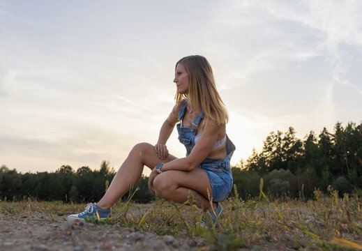 Attractive woman in denim overalls posing in nature during beautiful sunset, side view lifestyle portrait