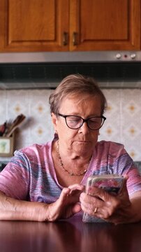 Senior woman with glasses sitting at a kitchen table while scrolling and typing on her smartphone, staying connected with family and friends through social media and technology