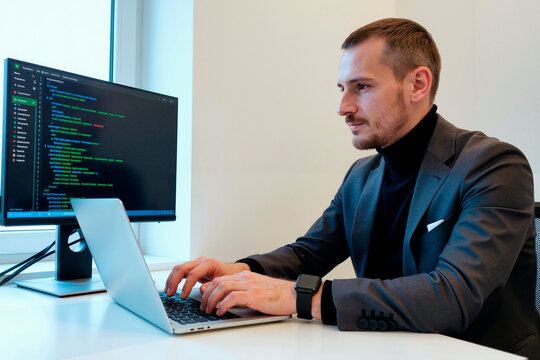 Medium shot of Caucasian young adult businessman coding on laptop beside software interface monitor, developing digital product. Ideal for IT services, programming, cybersecurity