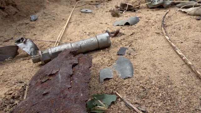 Stamped marking on a metal component of a KOBE 3B30 dual-purpose cluster submunition on sand, with an excavated pit of collected ordnance remains behind it