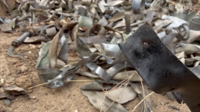 Heap of components from detonated KOBE 3B30 dual-purpose cluster submunitions: curled metal petals, casing fragments and faded fabric stabilizer ribbons on sandy ground