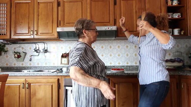 Joyful family moment with a happy senior mother and her adult daughter dancing and singing together while cooking in their home kitchen, sharing a beautiful moment of love and fun