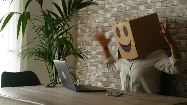 Happy businesswoman wearing a cardboard box with a smiley face on her head celebrating an achievement while working on her laptop computer, showing excitement and satisfaction