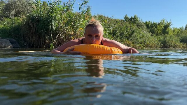 Mature woman floats on an orange inflatable ring in a calm river beside reed beds, spreading her arms and looking into camera in warm late summer light