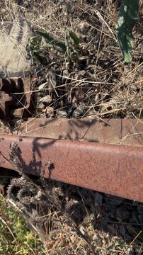 Rusty disused railway track overgrown with dried thistles and grass, from a close up of corroded rail bolts to the line receding past catenary masts under blue sky