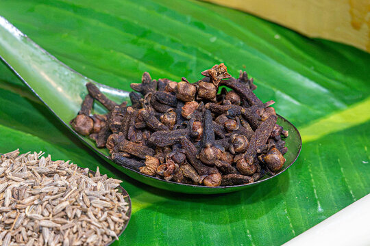 Close up of dried cloves in a spoon on a green banana leaf with cumin seeds