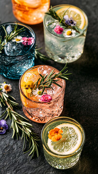 Colorful cocktail glasses garnished with rosemary, citrus and edible flowers arranged on a dark stone surface. Elegant botanical beverage display.