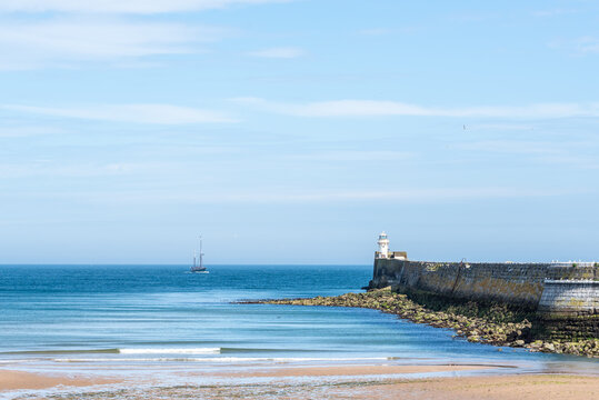 A calm North Sea scene along the Scottish coast, featuring a lighthouse with a green navigation light on a stone pier, a distant sailing vessel and gentle waves reaching the sandy shoreline under clea