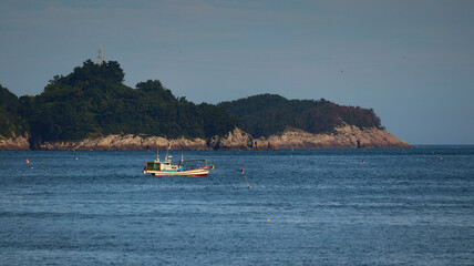 Small Fishing Boat Floating on Calm Blue Ocean Near Forested Island Coastline with Rocky Cliffs © L.Z.C Photographer