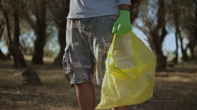 Close-up of environmental volunteer collecting plastic waste in wooded area using a yellow garbage bag and protective gloves. Forest cleanup, pollution prevention and nature conservation concept. 4K.