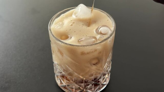 Cold dark fizzy drink is poured into a clear glass filled with ice cubes, creating bubbles and foam against a simple gray background. Close up.