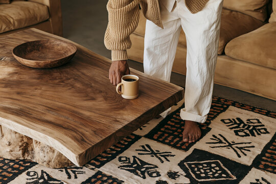 Man in ribbed knit sweater and slim cotton trousers placing coffee mug on live-edge coffee table in an afro boho living room with patterned rug