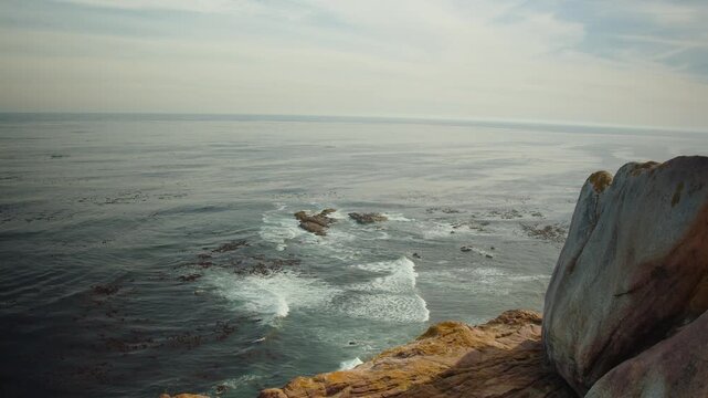 Rocky ocean coastline meets rolling waves beneath soft cloudy sky. Dark sea water surrounds offshore rocks while white surf traces shore, creating tranquil coastal nature background view.